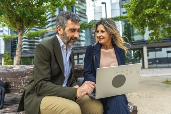 Mature business professionals, man and woman, discussing project ideas and networking on a park bench with a laptop in an urban setting, smiling and collaborating outdoors