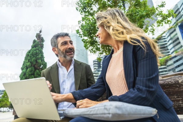 Two business colleagues engaged in a discussion while working on a laptop, sitting together on a bench in a bright urban park with modern buildings in the background