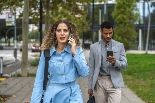 Businesswoman talking on her mobile phone while walking on a city sidewalk with a male colleague using his smartphone, both commuting and connecting in the modern urban environment