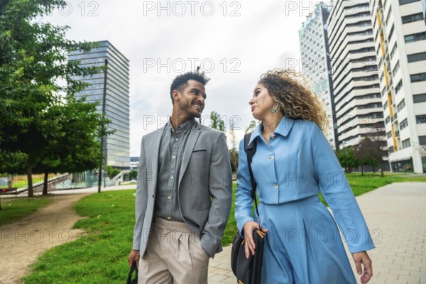 Two diverse business professionals walking along a path in a contemporary urban park with modern buildings, engaging in conversation while collaborating outdoors