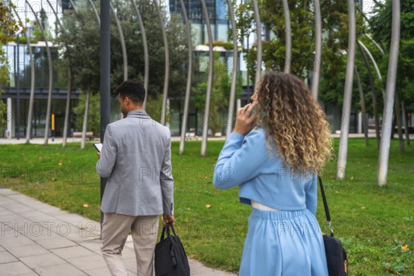Two diverse business colleagues, a man texting on his smartphone and a woman conversing on hers, walking outdoors in a urban park setting during a workday