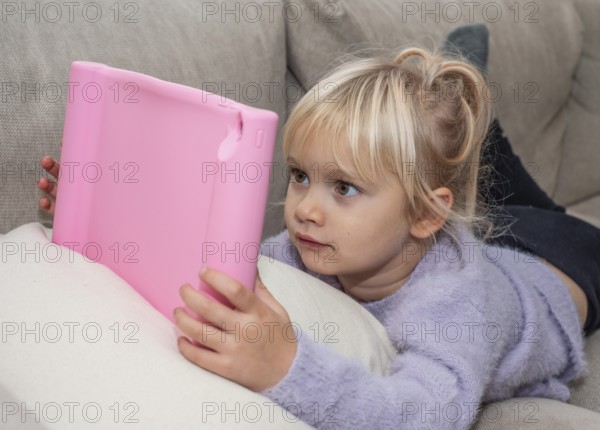Portrait of a blonde girl, 4 years old, lying down reading a tablet in Ystad, Skåne County, Sweden, Scandinavia