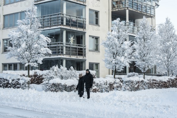 A couple walks on a snow-covered street in front of apartment buildings and snow-laden trees in Ystad, Skåne County, Sweden, Scandinavia
