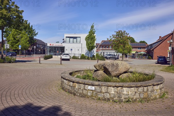 Vörden village square, fountain, commercial building, general architecture, Landessparkasse zu Oldenburg, parking lot with cars, paving stone walkway, barrier bollards, roads, trees, blue sky, cirrus clouds, Koppelstraße junction with Osnabrücker Straße, Vörden district, Neuenkirchen-Vörden, district of Vechta, Lower Saxony, Germany