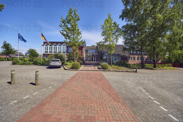 Town hall of the municipality of Neuenkirchen-Vörden, commercial building, modern architecture, parking lot with car, barrier bollard, flagpoles with European flag and German flag, paving brick walkway, concrete paving street, trees, blue sky, cirrus clouds, Küsterstraße, Neuenkirchen district, Neuenkirchen-Vörden, district of Vechta, Lower Saxony, Germany