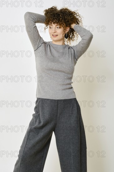 A woman stands with her hands behind her head, showcasing a cozy grey long-sleeve top paired with wide grey pants. The neutral backdrop enhances her fashionable look