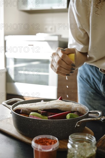 Close-up view of female hand squeezing lemon over hake fillet and vegetables in a baking pan