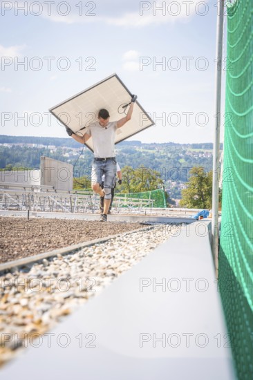 Man carrying a solar panel across a building site with a view and green fence, new park and ride parking garage Calw Heumaden on the new Hermann Hessebahn, Calw, Germany