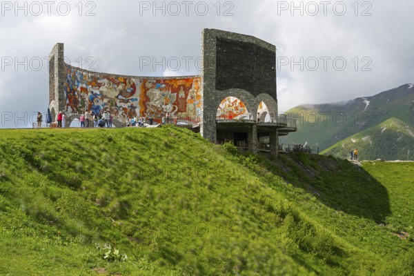 Grassy hill with painted wall, people enjoying the view, Georgian-Russian Friendship Memorial, observation deck, between Gudauri and the Cross Pass, Mtskheta-Mtianeti region, Georgian Military Route, High Caucasus, Georgia