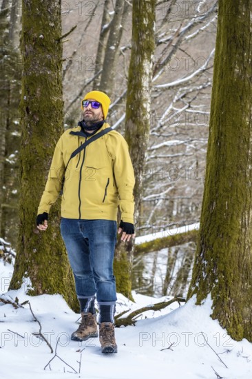 Man wearing a yellow winter jacket, jeans, and hiking boots exploring a snowy forest landscape, looking up at the trees while enjoying the peaceful nature and cold weather conditions