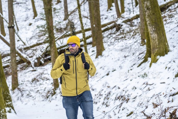 Man in yellow beanie and green jacket with backpack walking through snowy woodland, enjoying a winter trek along a quiet trail amid trees and serene, frosted landscape