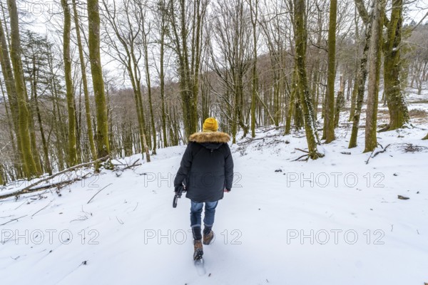 Person walking uphill in a winter forest, carrying a camera, wearing warm clothing with a yellow beanie and a hooded jacket, surrounded by bare trees during a cold day