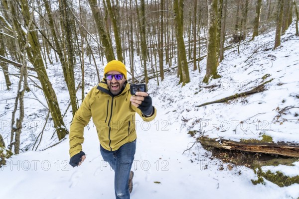 Happy man in a yellow beanie and green jacket smiling at his action camera while vlogging on a snowy forest hiking trail, capturing winter adventure and outdoor fun