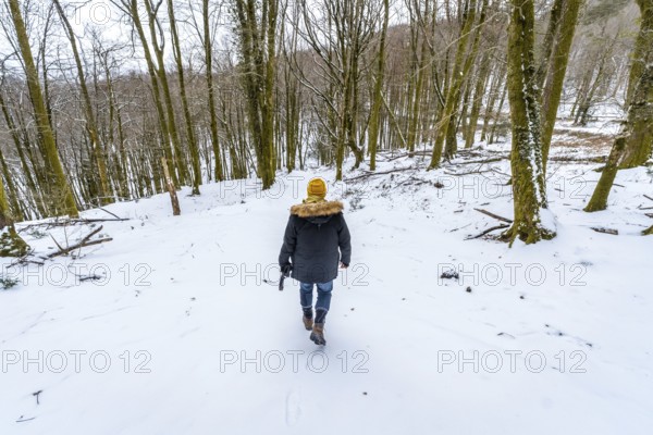 Man in a winter coat and yellow hat walking away into a snow covered forest, exploring and enjoying the cold outdoor nature during a peaceful and adventurous winter hike