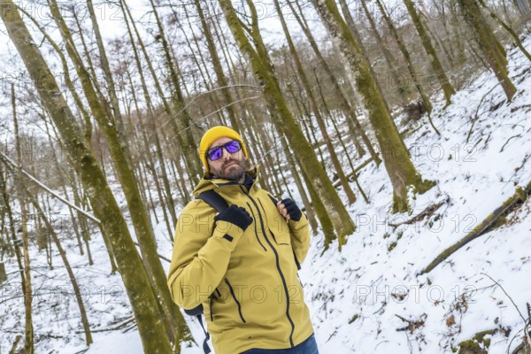 Man wearing a yellow beanie and jacket, sunglasses, and backpack, standing confident and looking at the camera while trekking through a beautiful snow covered forest during winter