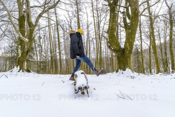 Man wearing winter clothing and a yellow beanie balancing on a snow covered log, enjoying the cold weather and adventure during a winter hike in a bare forest
