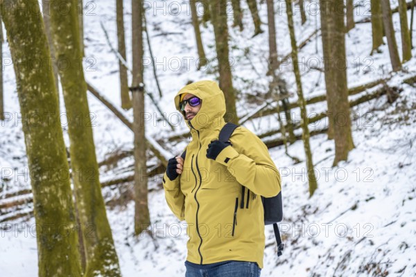 Man in yellow hooded jacket and backpack standing in snowy forest, exploring winter trails and enjoying a peaceful outdoor adventure amid silent trees and cold landscape
