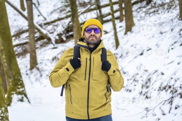 Man in yellow jacket and beanie with sunglasses and backpack hiking through a snow covered forest in winter, enjoying a peaceful, frosty outdoor adventure and scenic landscape