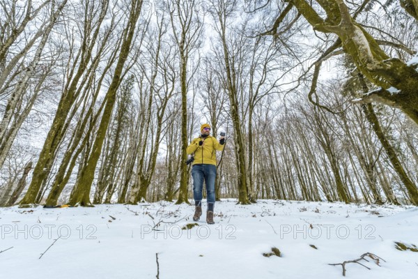 Man in a yellow jacket and beanie enjoying a winter adventure, holding a camera and phone while standing amidst tall, bare trees in a snow covered forest