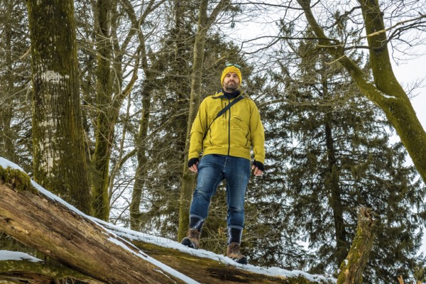 Man in yellow jacket and beanie standing on a fallen log in a snowy forest, confidently taking in a peaceful winter hiking scene that evokes adventure and solitude