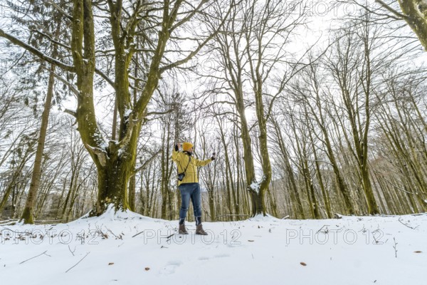 Man in yellow jacket and beanie enjoying a solitary winter hike, standing in a snow covered forest surrounded by tall bare trees, looking up and reaching arms out