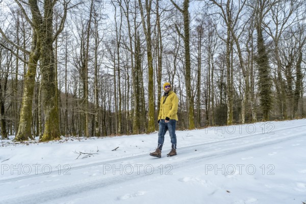 Man in a yellow jacket, beanie, and sunglasses exploring a snow covered forest path, enjoying the cold weather and serene natural environment during adventure travel