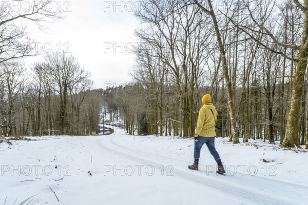 Person with yellow jacket and beanie beanie walking down a snow covered road winding through a bare winter forest, surrounded by trees on a cold day, enjoying nature and solitude