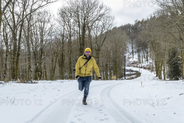 Cheerful man in a yellow jacket and hat joyfully running on a snow covered forest path, expressing happiness and freedom during a winter adventure in nature