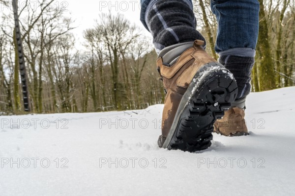 Person walking through a winter forest landscape wearing brown hiking boots, their soles visible tracking in fresh white snow while exploring the outdoors
