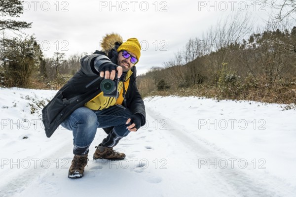 Man in winter gear with a yellow hat and sunglasses crouching on a snowy trail, aiming a camera to capture a scenic forest moment during a cold outdoor hike