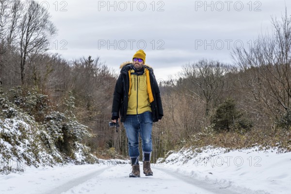 Man in warm winter attire and sunglasses walking on a snowy road through a woodland landscape, holding a compact camera and enjoying outdoor adventure in cold weather