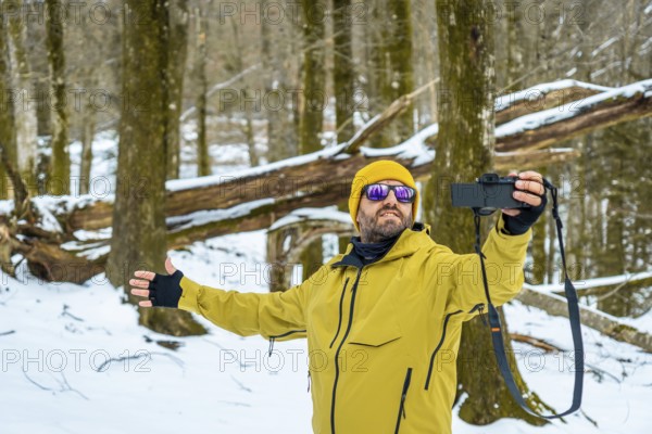 Man wearing a yellow jacket and beanie capturing moments with his camera in a snow covered forest, documenting his winter adventure and sharing his travel experience