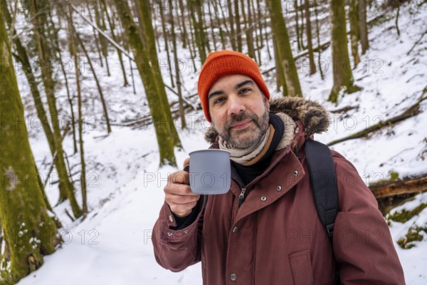 Man with an orange beanie and a winter coat holding a mug of a hot beverage, finding warmth and relaxation during a winter outdoor activity in a snow covered forest
