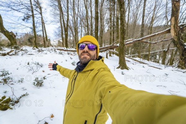 Man posing for a selfie during a winter hike, exploring a snowy forest landscape with bare trees and fallen branches, enjoying the quiet nature and cold weather travel