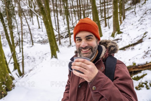 Bearded man smiling in a winter hat and jacket, holding a steaming mug while standing in a snow covered forest on a peaceful outdoor hiking adventure