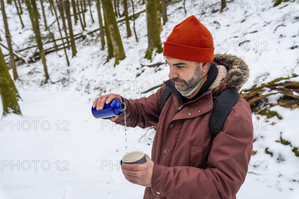 Man wearing an orange beanie and winter jacket poring a hot drink from a blue thermos into a mug, taking a break during a winter hike in a snow covered forest