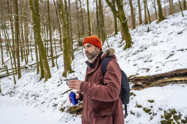 Adventurous man in a beanie and jacket pauses on a snowy forest trail with a backpack, sipping a warm drink from a thermos and enjoying a peaceful winter hike