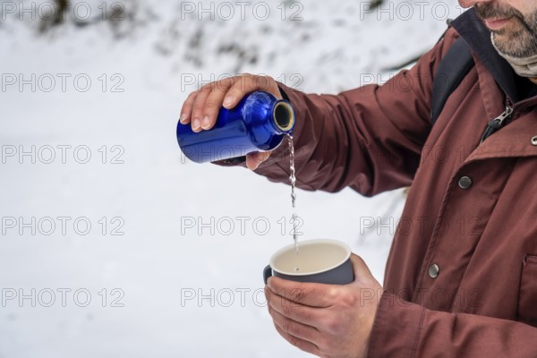 Man wearing a winter jacket pouring a hot drink from a blue thermos bottle into a cup, staying refreshed and hydrated while outdoors in a cold snowy environment