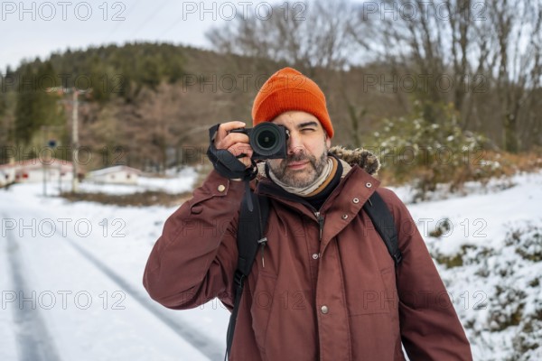 Man in winter clothing and an orange beanie holding a mirrorless camera, capturing nature and winter landscapes on a snowy path during a cold weather adventure
