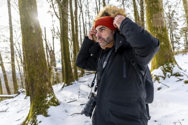 Man photographer adjusting his jacket or coat hood with fur trim while wearing a beanie and scarf outdoors in a snowy forest during winter with trees and sunlight