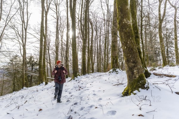 Man wearing winter clothing and a backpack walking on a snow covered path through a barren forest, enjoying the serene outdoor landscape and cold weather