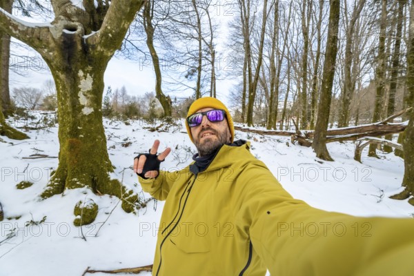 Man smiling at the camera and making a peace sign gesture, wearing a yellow jacket, beanie, and purple reflective sunglasses in a snow covered forest during winter