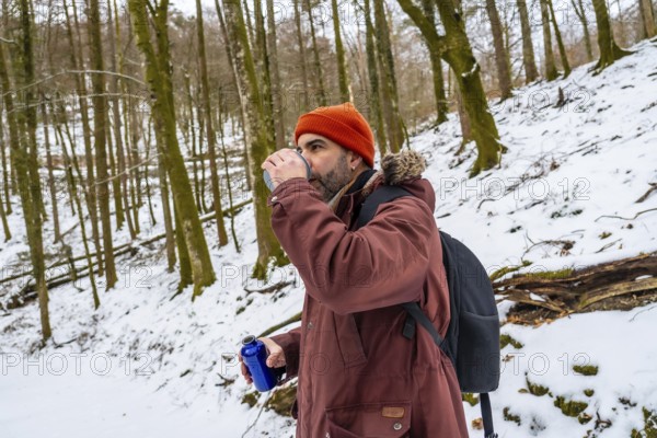 Man in orange beanie and winter jacket drinks from a reusable bottle on a snowy forest trail during a winter hike, pausing to hydrate and enjoy the peaceful outdoor adventure