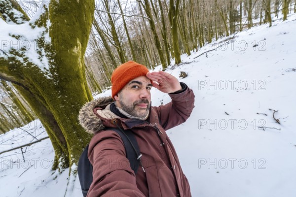 Man posing for a selfie in a snowy forest during winter, looking out with his hand shading his eyes, wearing an orange beanie and a warm jacket for outdoor adventure