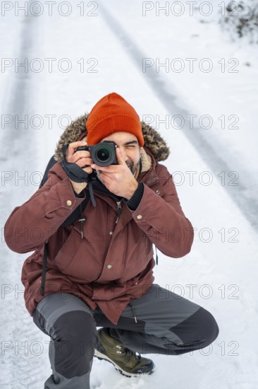Photographer wearing an orange beanie and parka kneeling in the snow, capturing images with a digital camera during a cold winter day in a natural outdoor environment