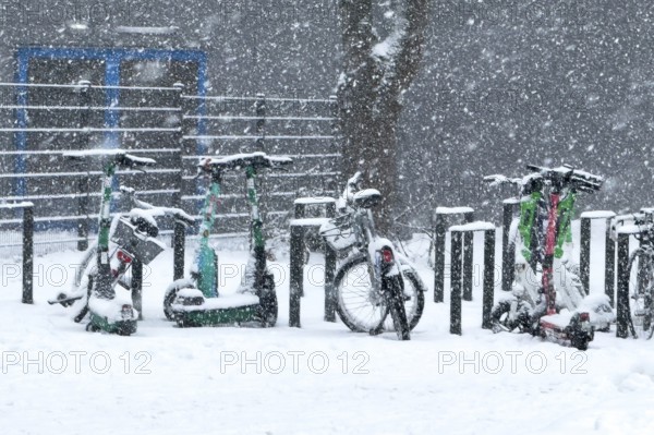 Tailored parked e-scooters and bicycles in winter during snowfall with snowflakes in the picture, Germany