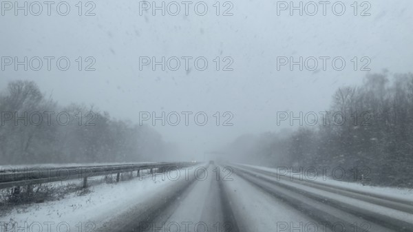 View through the windshield of a passenger car driving through light snow on an empty, partially tailored motorway without traffic with lanes covered by snow, Germany