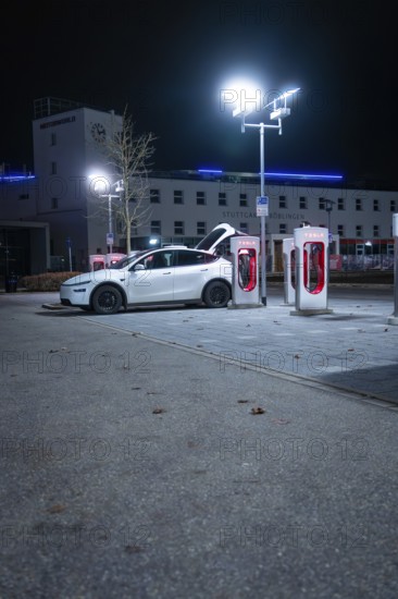 Parking lot at night with white Tesla and charging stations under city lights, Tesla Supercharger, Böblingen, Germany