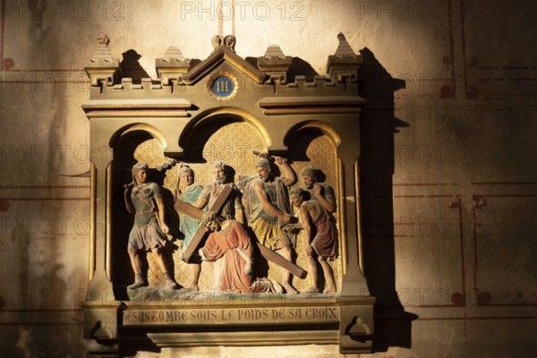 A carved relief depicts a religious scene. Jesus falls while carrying the cross. Roman soldiers and other figures surround him in a church. The text reads, JESUS TOMBE SOUS LE POIDS DE SA CROIX