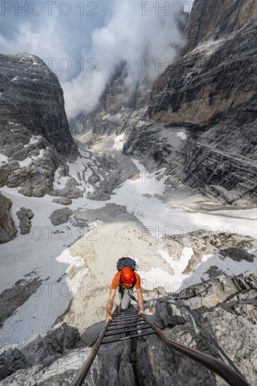 Mountaineer climbs a ladder in steep rocky terrain, Via Ferrata Oliva Detassis via ferrata, view of mountain basin with steep rock walls covered in clouds, Brenta Mountains, Trentino, Italy